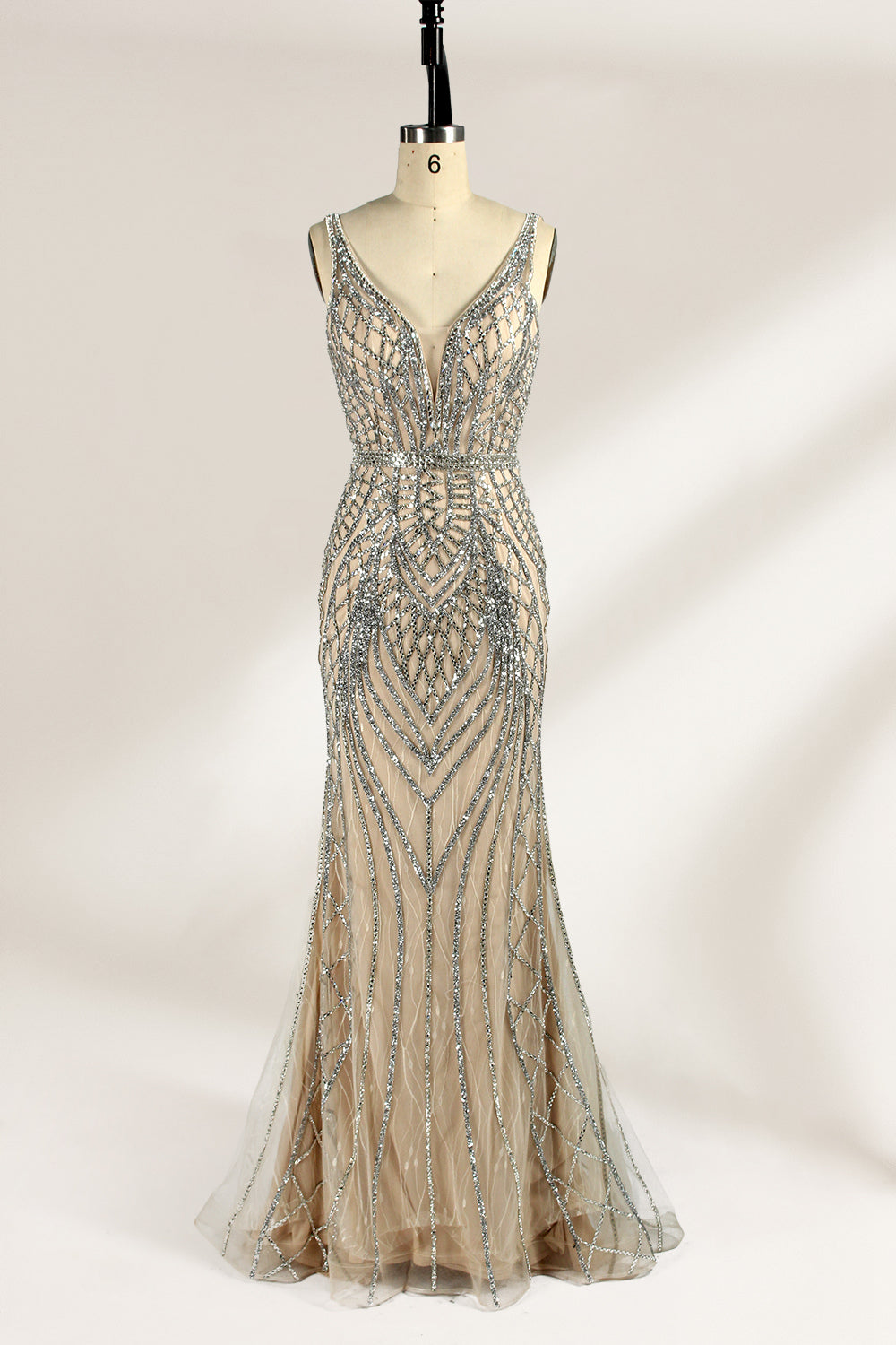 Against a light - hued backdrop, a mannequin displays a nude - toned tulle gown featuring silver beaded embellishments. It has thin straps, a deep V - neckline, and a mermaid - shaped skirt. The detailed beadwork weaves sophistication, perfect for formal dinners or weddings, where the beaded elegance complements the occasion.1