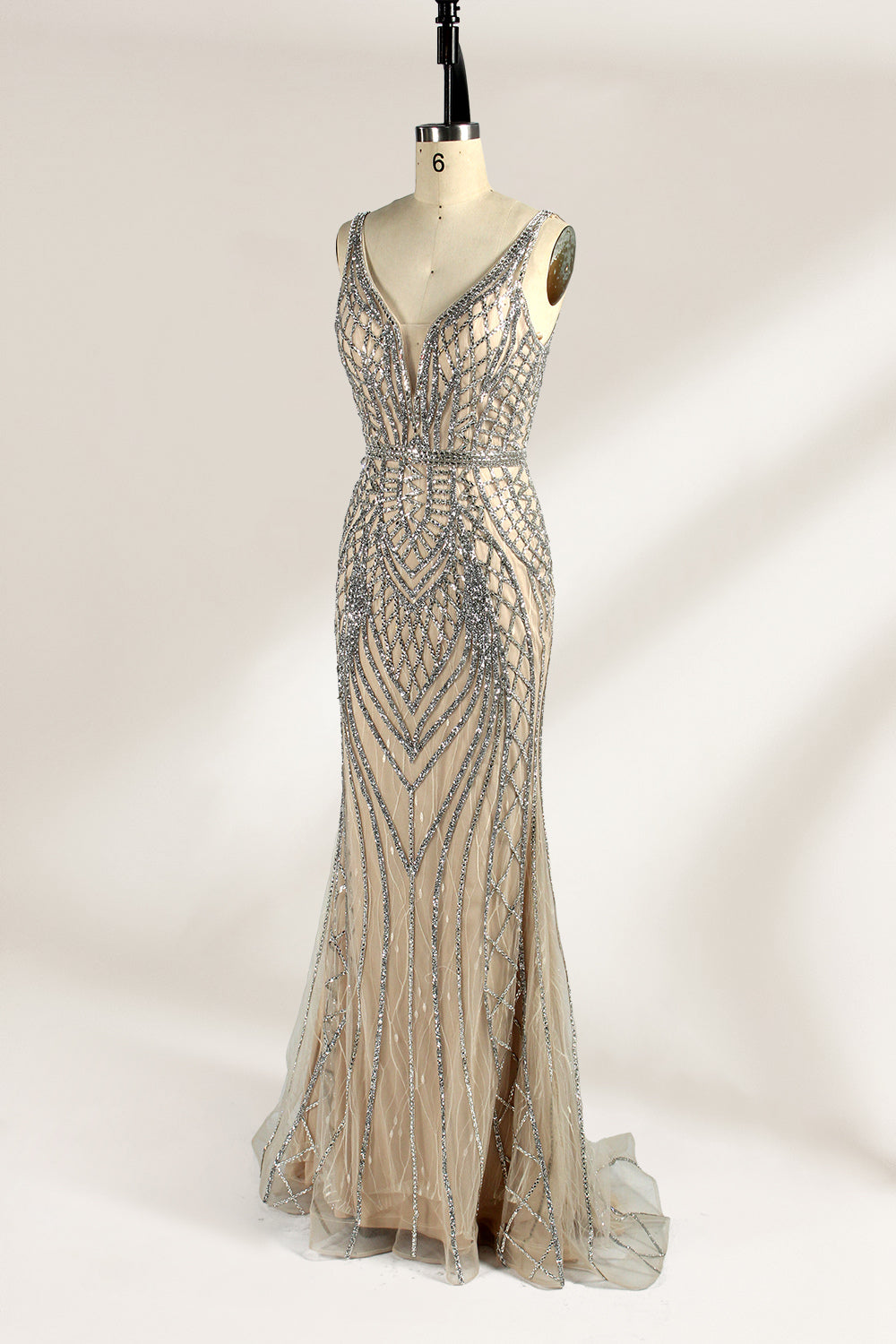 Against a light - hued backdrop, a mannequin displays a nude - toned tulle gown featuring silver beaded embellishments. It has thin straps, a deep V - neckline, and a mermaid - shaped skirt. The detailed beadwork weaves sophistication, perfect for formal dinners or weddings, where the beaded elegance complements the occasion.4