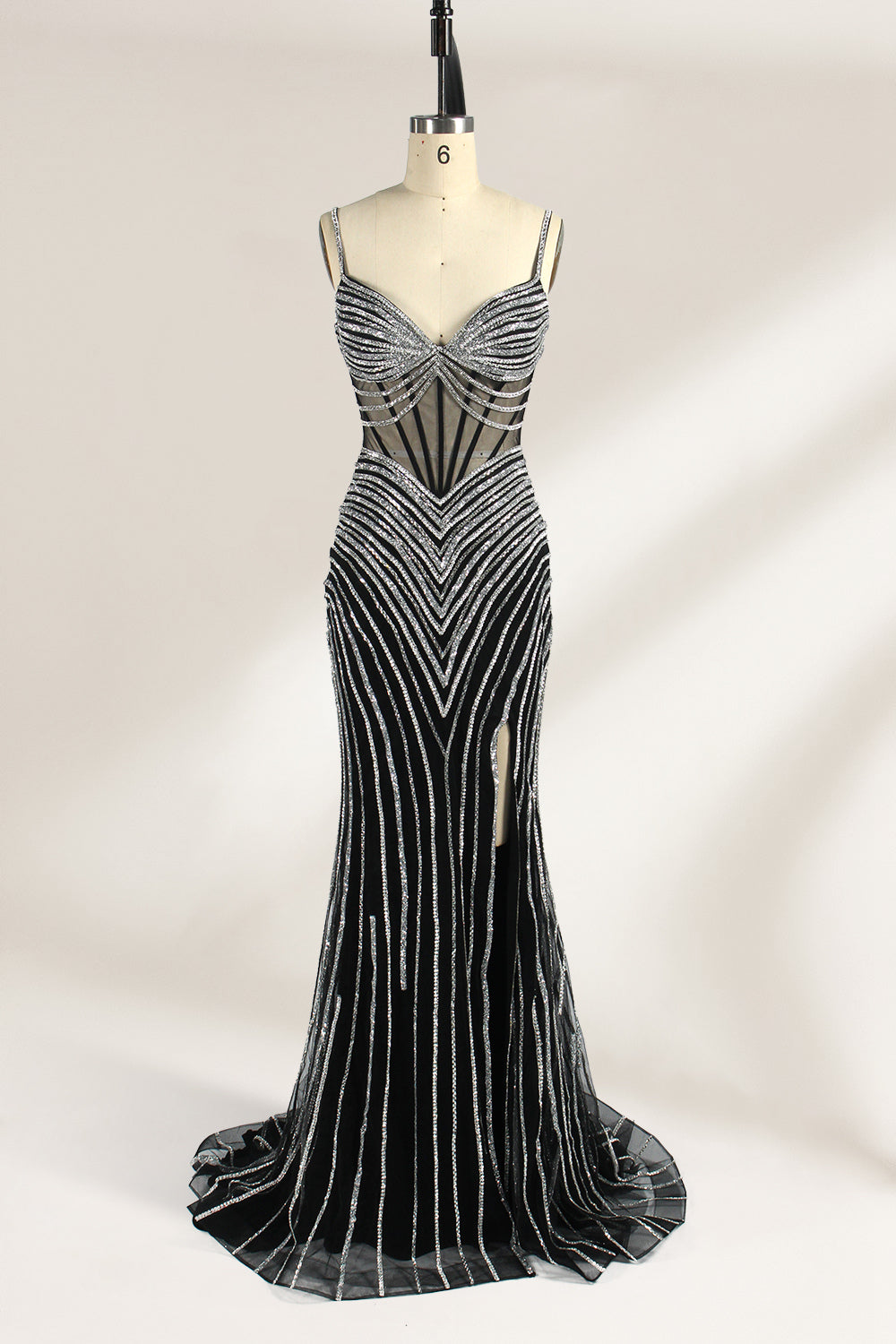 A mannequin numbered “6” displays a black tulle gown against a light backdrop. It features thin straps, a deep V - neckline, and a mermaid silhouette. The intricate silver bead patterns, sheer bodice with vertical accents, and high side slit add glamour, perfect for formal galas.1