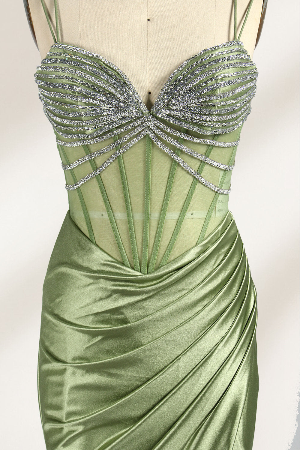 A mannequin presents a green satin gown with a bejeweled, sheer bodice against a light backdrop. It features thin straps, a sweetheart neckline, and a mermaid - shaped skirt. The sparkling embellishments and smooth fabric exude elegance, perfect for formal galas.6