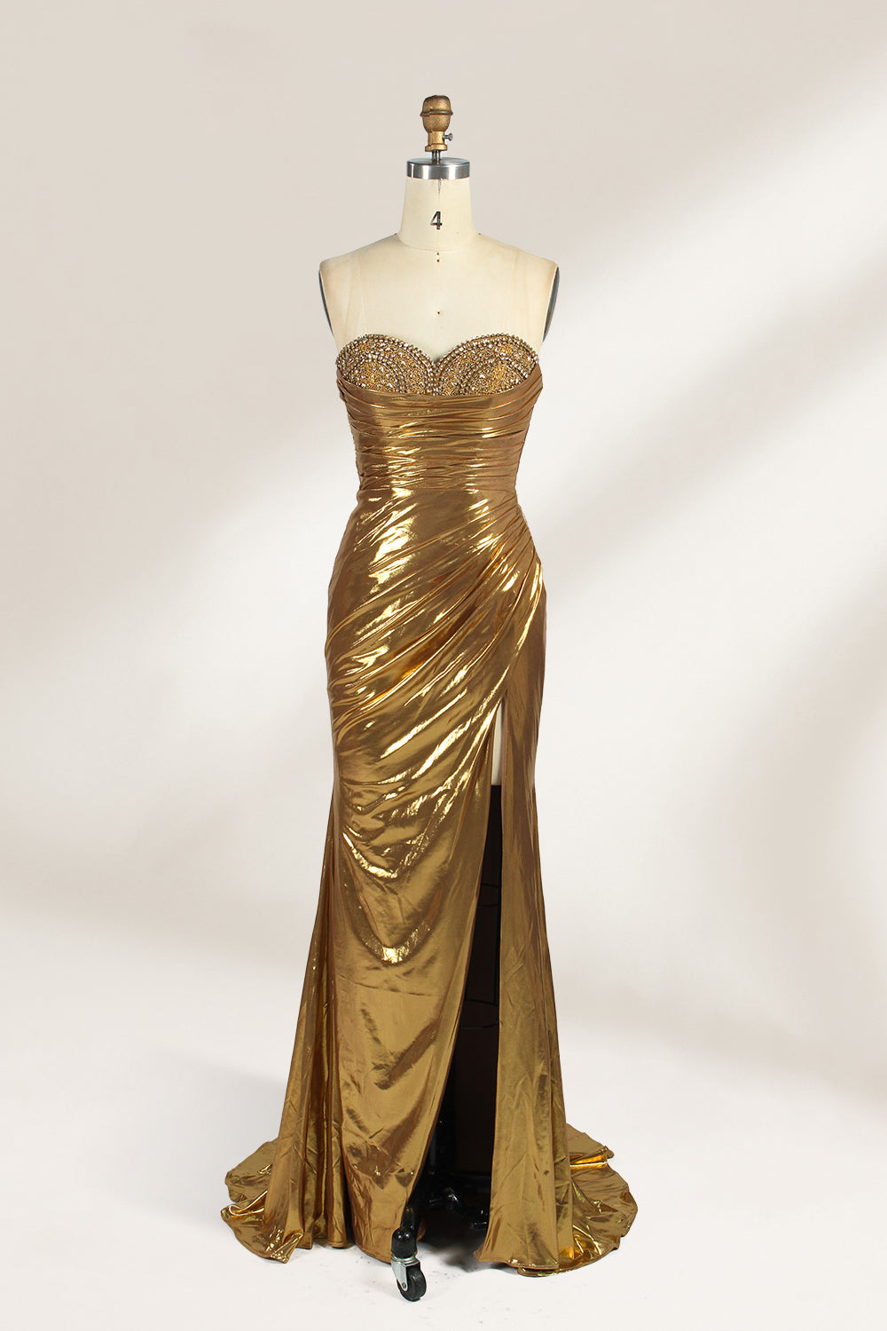 A golden strapless evening gown is displayed on a mannequin. The bodice is adorned with intricate beadwork, and the skirt, made of a shiny metallic fabric, features a high side slit and a small train, creating a glamorous and elegant look, perfect for formal events.1