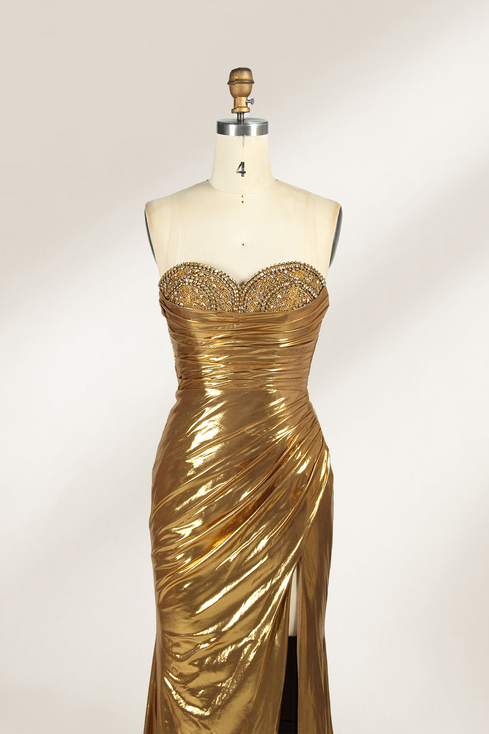 A golden strapless evening gown is displayed on a mannequin. The bodice is adorned with intricate beadwork, and the skirt, made of a shiny metallic fabric, features a high side slit and a small train, creating a glamorous and elegant look, perfect for formal events.4