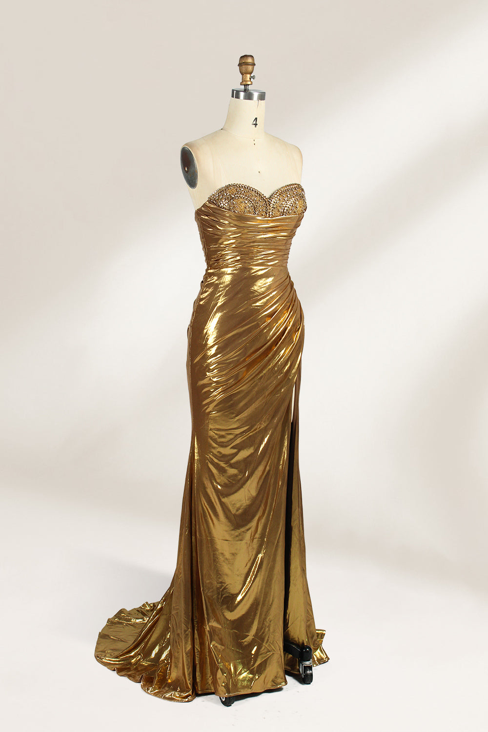 A golden strapless evening gown is displayed on a mannequin. The bodice is adorned with intricate beadwork, and the skirt, made of a shiny metallic fabric, features a high side slit and a small train, creating a glamorous and elegant look, perfect for formal events.3