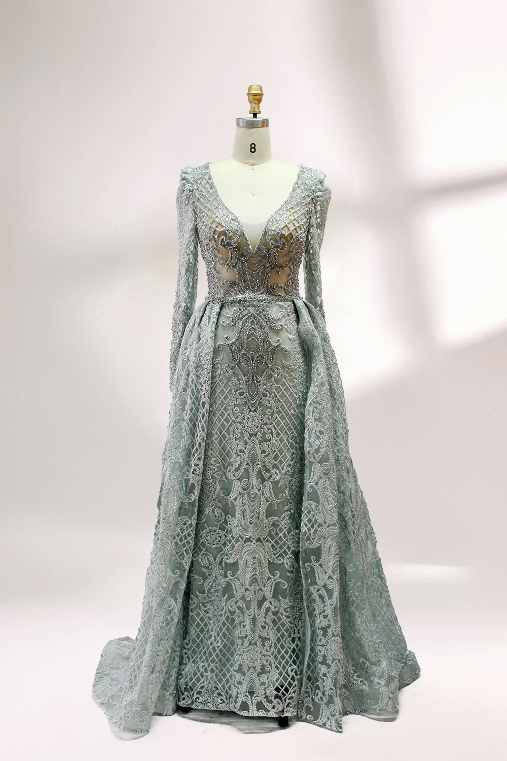 Displayed on a mannequin is a light - gray evening gown. It has a deep V - neckline, long sleeves, and intricate lace embroidery with a grid pattern. The elegant design gives off a luxurious and sophisticated vibe, ideal for formal occasions.1