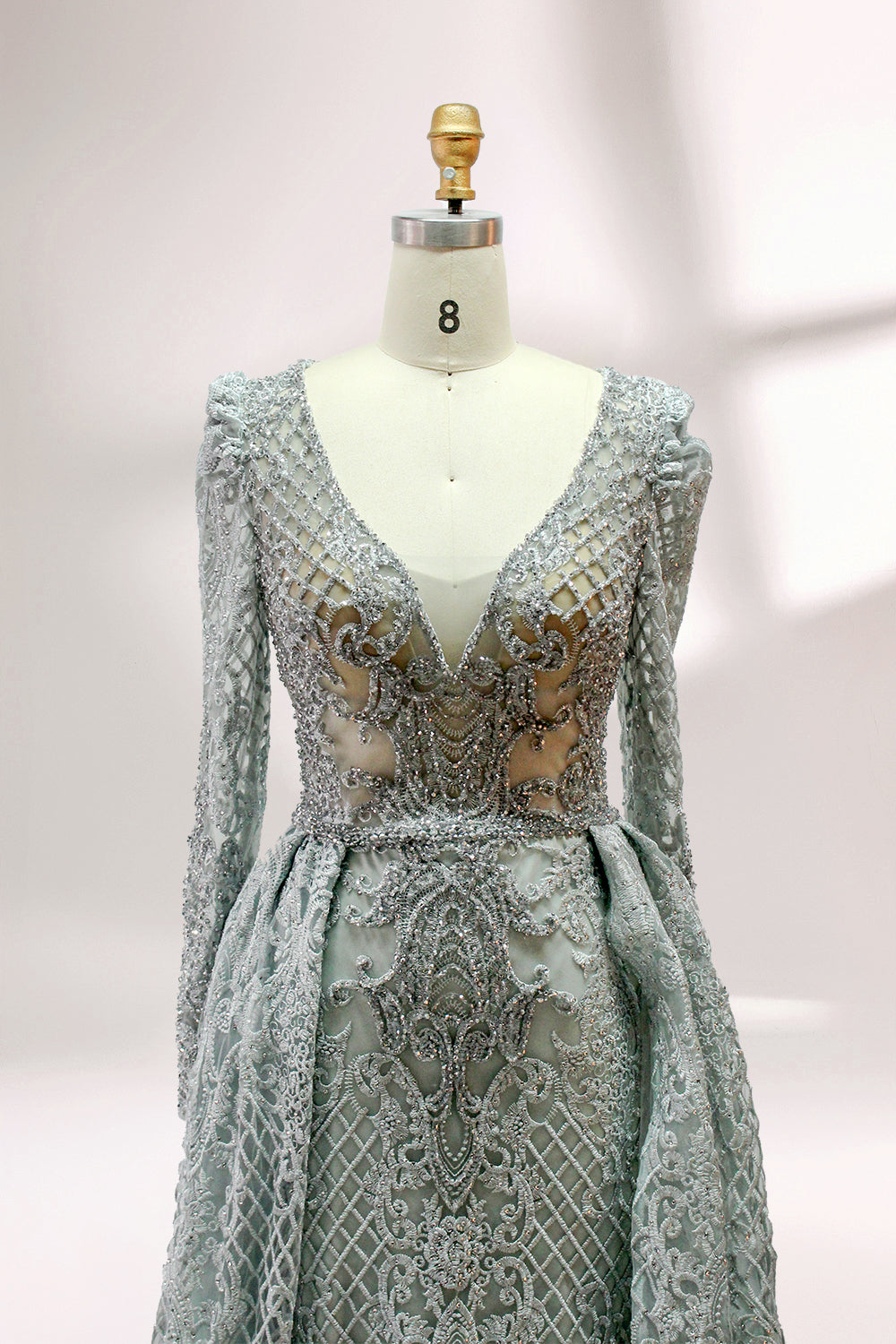 Displayed on a mannequin is a light - gray evening gown. It has a deep V - neckline, long sleeves, and intricate lace embroidery with a grid pattern. The elegant design gives off a luxurious and sophisticated vibe, ideal for formal occasions.8