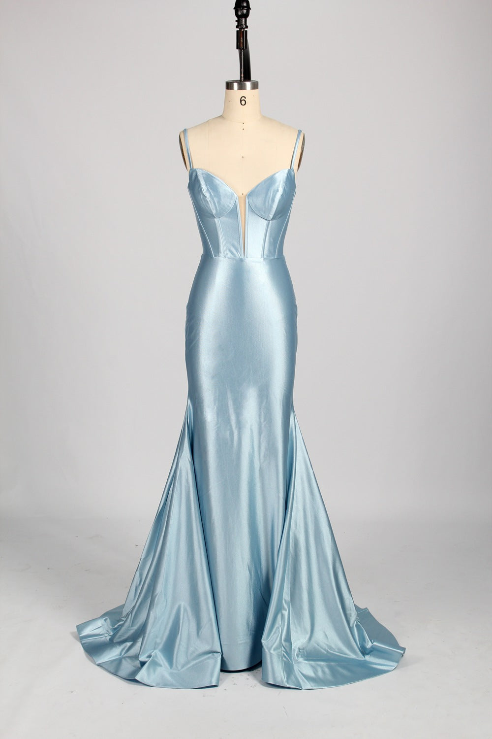 Photograph of a slender, fair-skinned woman with dark hair in an elegant updo, wearing a stunning, floor-length, light blue satin gown with a plunging neckline and thin spaghetti straps. The dress features a fitted bodice and a flowing skirt. She stands against a plain, light gray background, with soft lighting emphasizing the gown's sheen.9