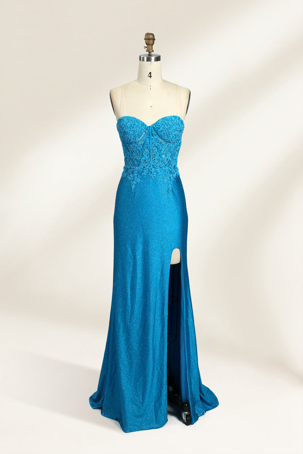Photograph of a mannequin dressed in a strapless, floor-length, vibrant blue evening gown. The gown features intricate lace detailing across the bodice, with a fitted, corset-style silhouette. The skirt has a high slit on the right side, revealing black high heels underneath. The background is a plain, light beige, emphasizing the gown's shimmering texture and elegant design. The mannequin stands on a wooden base, labeled "4" on its neck.01
