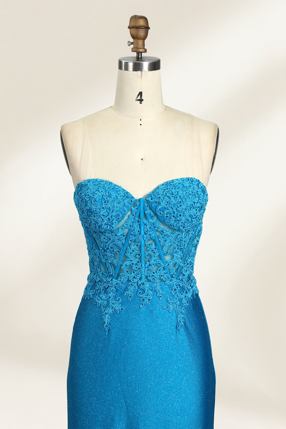 Photograph of a mannequin dressed in a strapless, floor-length, vibrant blue evening gown. The gown features intricate lace detailing across the bodice, with a fitted, corset-style silhouette. The skirt has a high slit on the right side, revealing black high heels underneath. The background is a plain, light beige, emphasizing the gown's shimmering texture and elegant design. The mannequin stands on a wooden base, labeled "4" on its neck.03