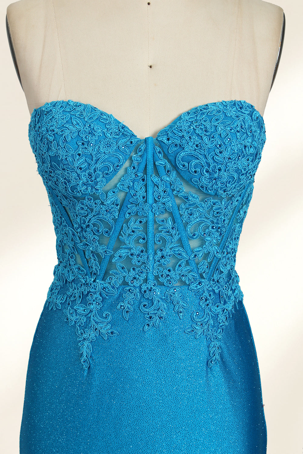 Photograph of a mannequin dressed in a strapless, floor-length, vibrant blue evening gown. The gown features intricate lace detailing across the bodice, with a fitted, corset-style silhouette. The skirt has a high slit on the right side, revealing black high heels underneath. The background is a plain, light beige, emphasizing the gown's shimmering texture and elegant design. The mannequin stands on a wooden base, labeled "4" on its neck.04