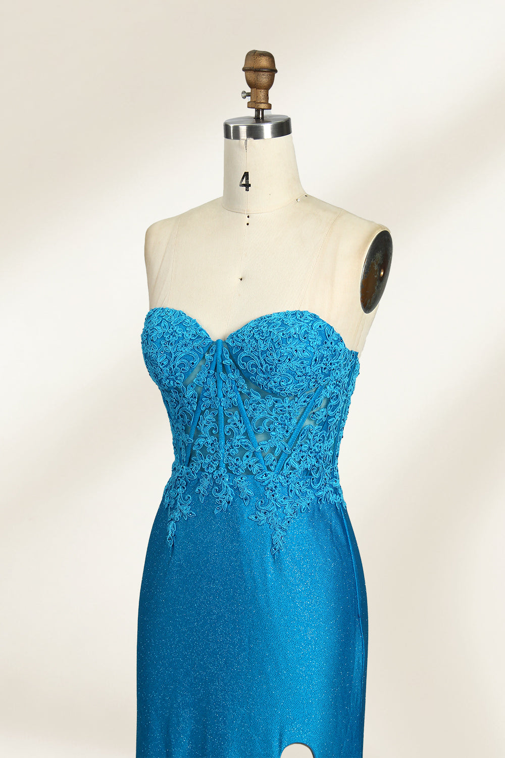 Photograph of a mannequin dressed in a strapless, floor-length, vibrant blue evening gown. The gown features intricate lace detailing across the bodice, with a fitted, corset-style silhouette. The skirt has a high slit on the right side, revealing black high heels underneath. The background is a plain, light beige, emphasizing the gown's shimmering texture and elegant design. The mannequin stands on a wooden base, labeled "4" on its neck.05