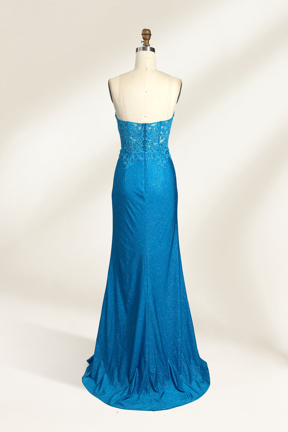 Photograph of a mannequin dressed in a strapless, floor-length, vibrant blue evening gown. The gown features intricate lace detailing across the bodice, with a fitted, corset-style silhouette. The skirt has a high slit on the right side, revealing black high heels underneath. The background is a plain, light beige, emphasizing the gown's shimmering texture and elegant design. The mannequin stands on a wooden base, labeled "4" on its neck.02