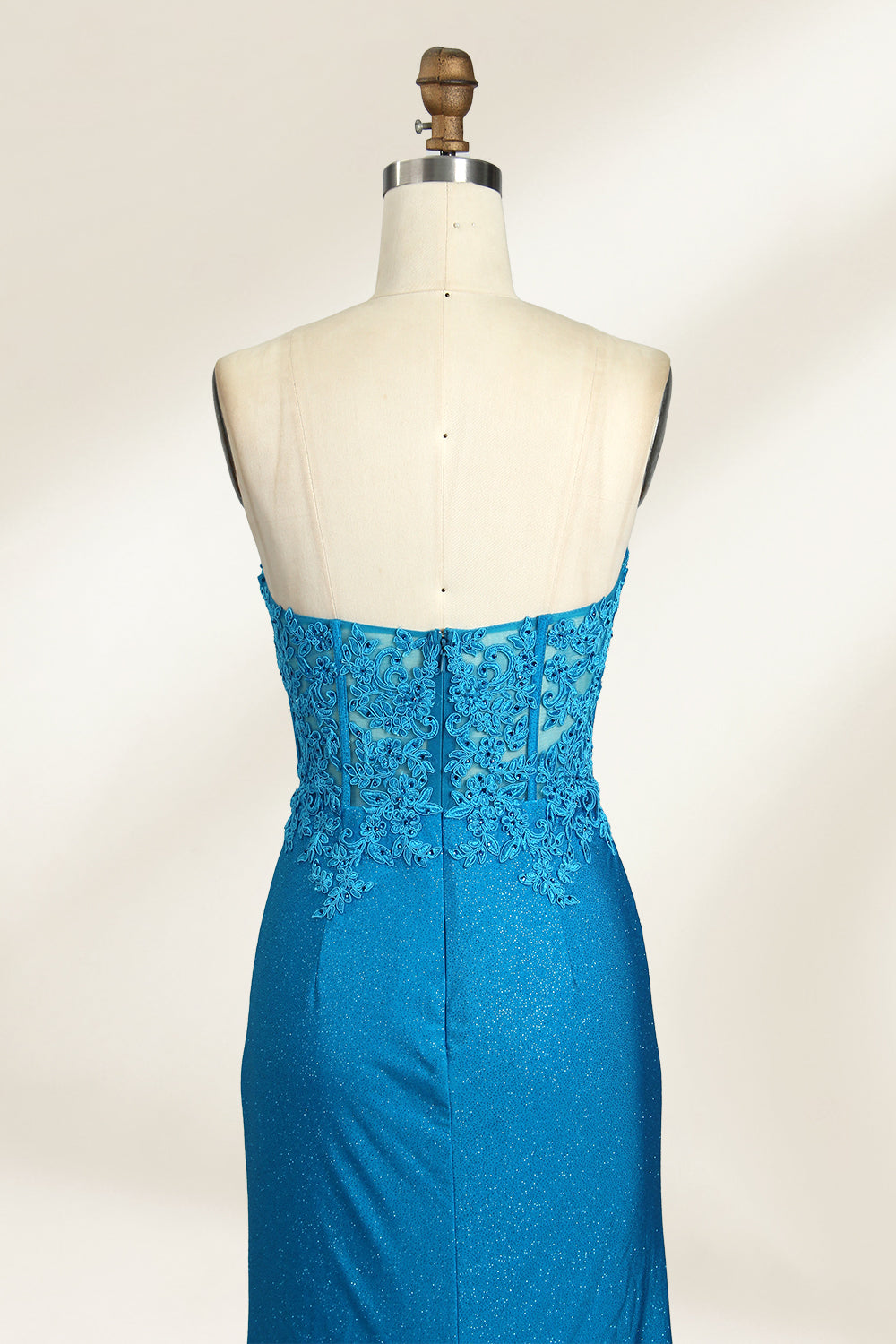 Photograph of a mannequin dressed in a strapless, floor-length, vibrant blue evening gown. The gown features intricate lace detailing across the bodice, with a fitted, corset-style silhouette. The skirt has a high slit on the right side, revealing black high heels underneath. The background is a plain, light beige, emphasizing the gown's shimmering texture and elegant design. The mannequin stands on a wooden base, labeled "4" on its neck.06