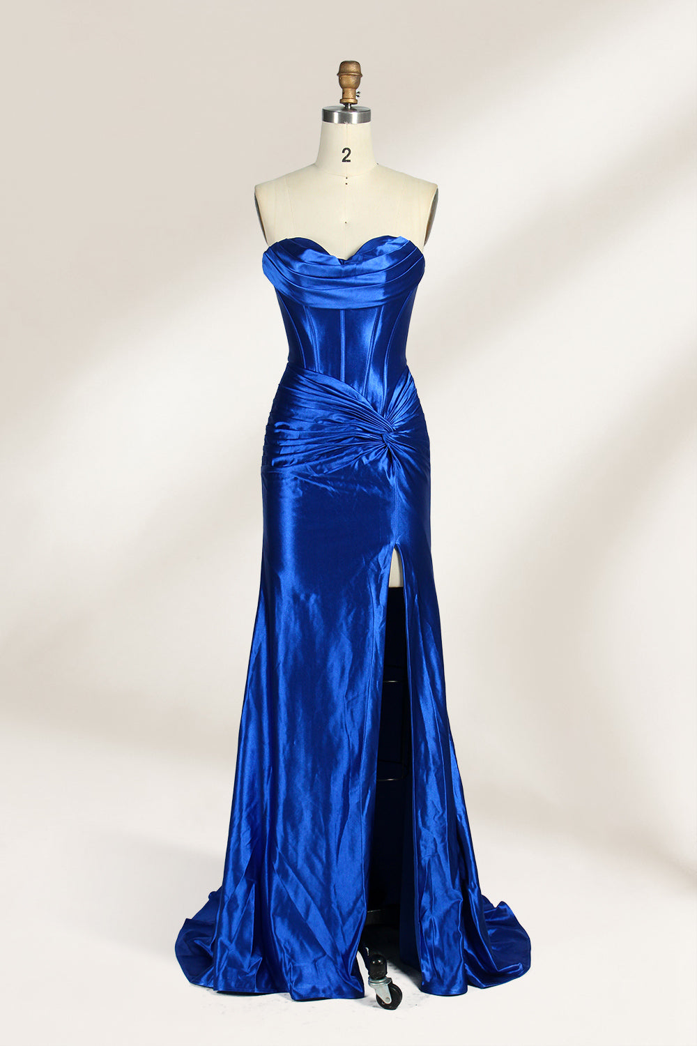 A mannequin showcases a strapless, blush - pink satin gown against a light backdrop. It features a ruched bodice with a draped neckline and a twisted detail at the waist, along with a mermaid silhouette and a side slit. The smooth satin fabric exudes elegance, perfect for formal events like galas.1