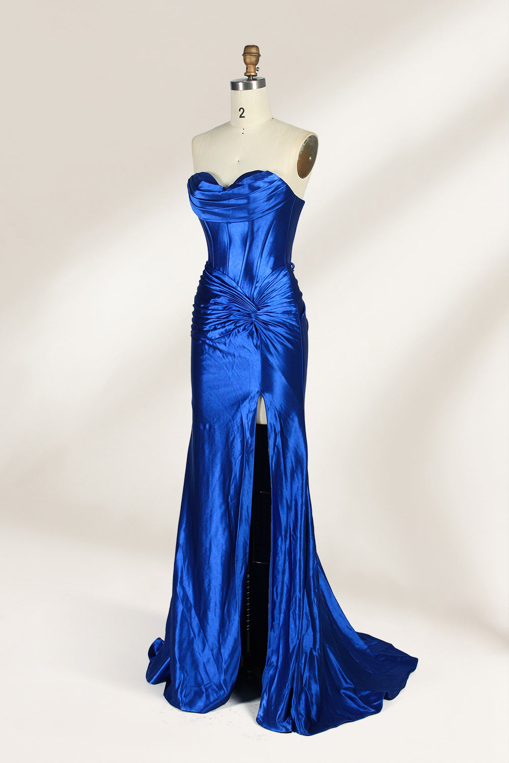A mannequin showcases a strapless, blush - pink satin gown against a light backdrop. It features a ruched bodice with a draped neckline and a twisted detail at the waist, along with a mermaid silhouette and a side slit. The smooth satin fabric exudes elegance, perfect for formal events like galas.3