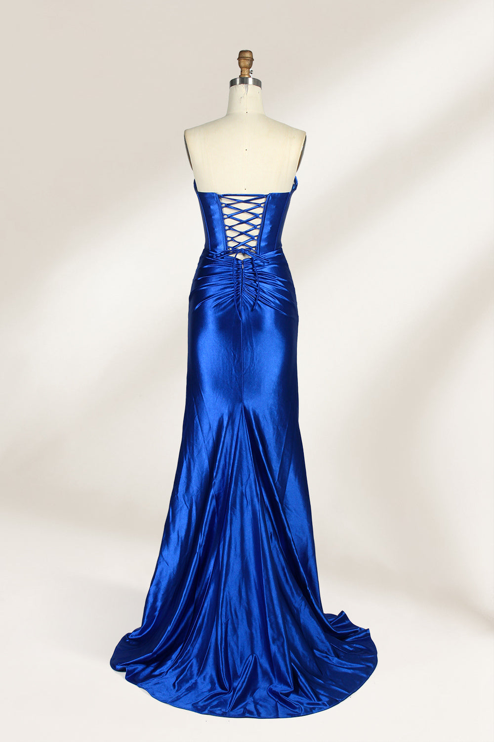 A mannequin showcases a strapless, blush - pink satin gown against a light backdrop. It features a ruched bodice with a draped neckline and a twisted detail at the waist, along with a mermaid silhouette and a side slit. The smooth satin fabric exudes elegance, perfect for formal events like galas.2