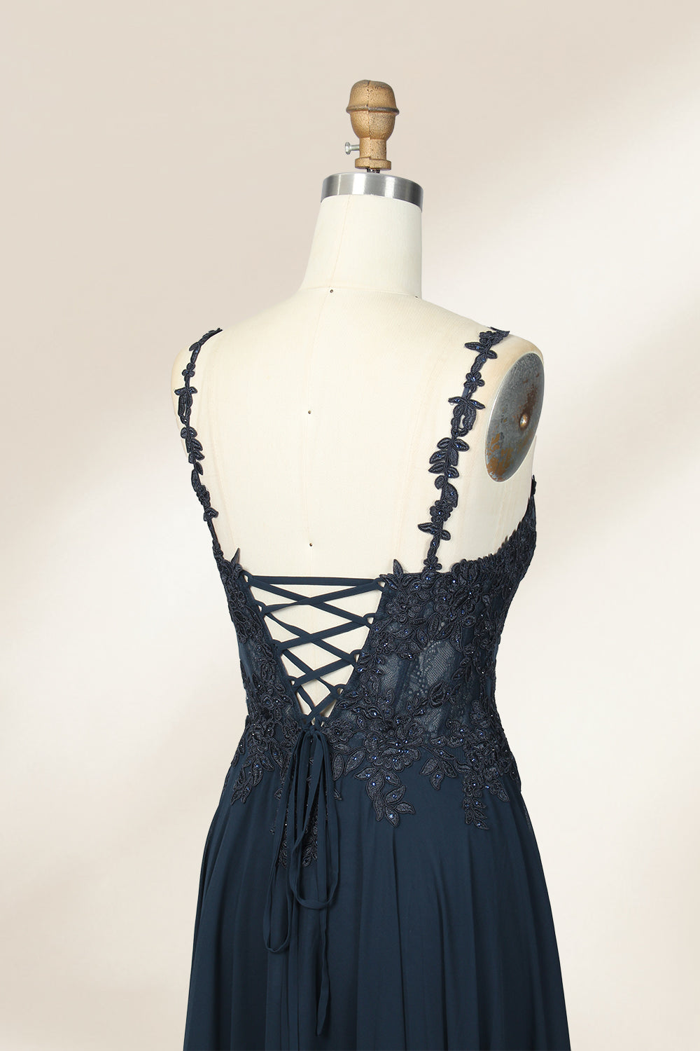 Photograph of a blue, floor-length, sleeveless gown with intricate lace detailing on the bodice and a subtle thigh-high slit, displayed on a mannequin. The gown has a fitted top and a flowing, soft fabric skirt. The background is plain beige.04