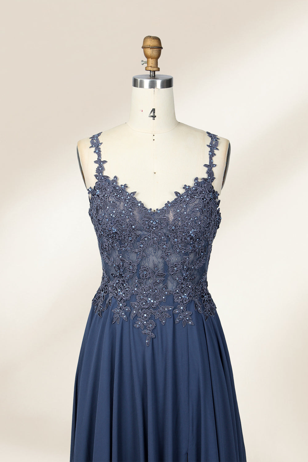 Photograph of a blue, floor-length, sleeveless gown with intricate lace detailing on the bodice and a subtle thigh-high slit, displayed on a mannequin. The gown has a fitted top and a flowing, soft fabric skirt. The background is plain beige.03