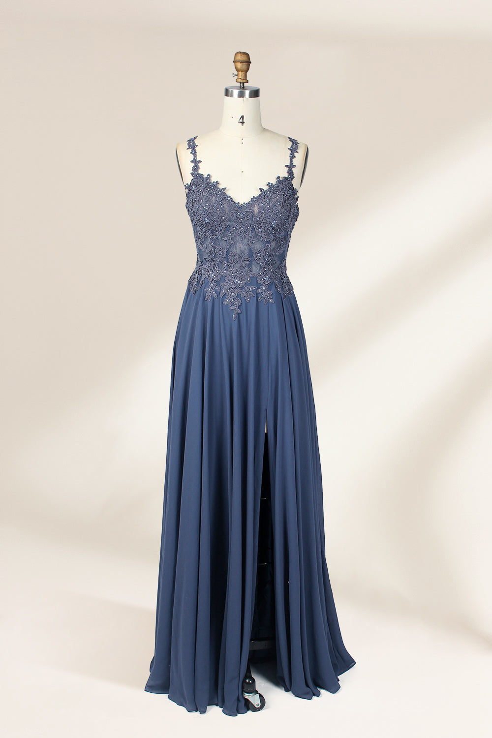 Photograph of a blue, floor-length, sleeveless gown with intricate lace detailing on the bodice and a subtle thigh-high slit, displayed on a mannequin. The gown has a fitted top and a flowing, soft fabric skirt. The background is plain beige.01