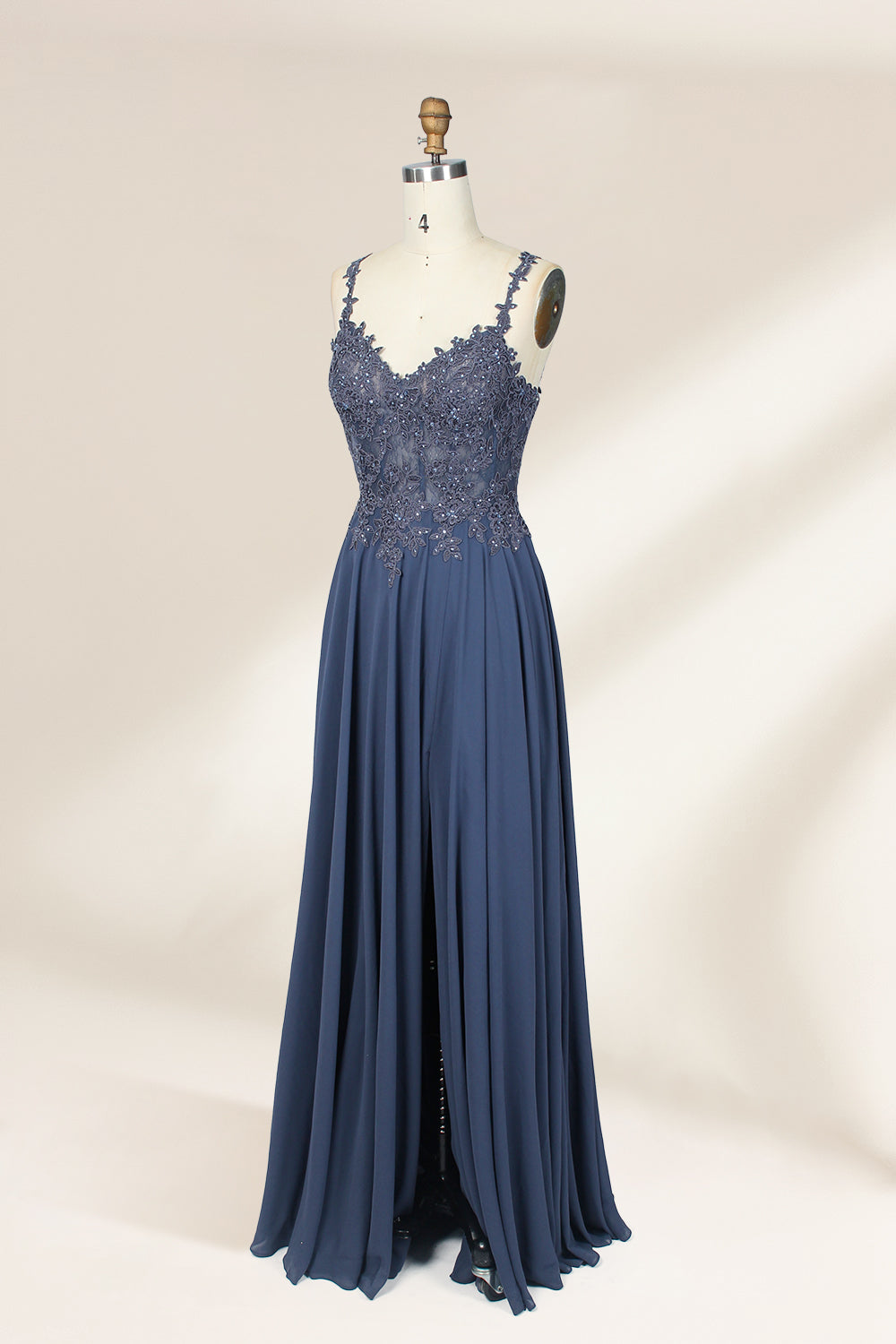 Photograph of a blue, floor-length, sleeveless gown with intricate lace detailing on the bodice and a subtle thigh-high slit, displayed on a mannequin. The gown has a fitted top and a flowing, soft fabric skirt. The background is plain beige.05
