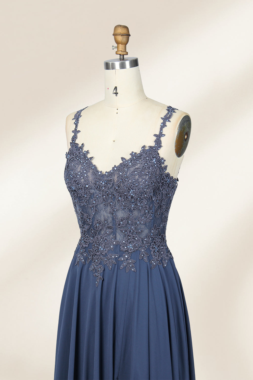 Photograph of a blue, floor-length, sleeveless gown with intricate lace detailing on the bodice and a subtle thigh-high slit, displayed on a mannequin. The gown has a fitted top and a flowing, soft fabric skirt. The background is plain beige.06