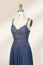 Photograph of a blue, floor-length, sleeveless gown with intricate lace detailing on the bodice and a subtle thigh-high slit, displayed on a mannequin. The gown has a fitted top and a flowing, soft fabric skirt. The background is plain beige.06