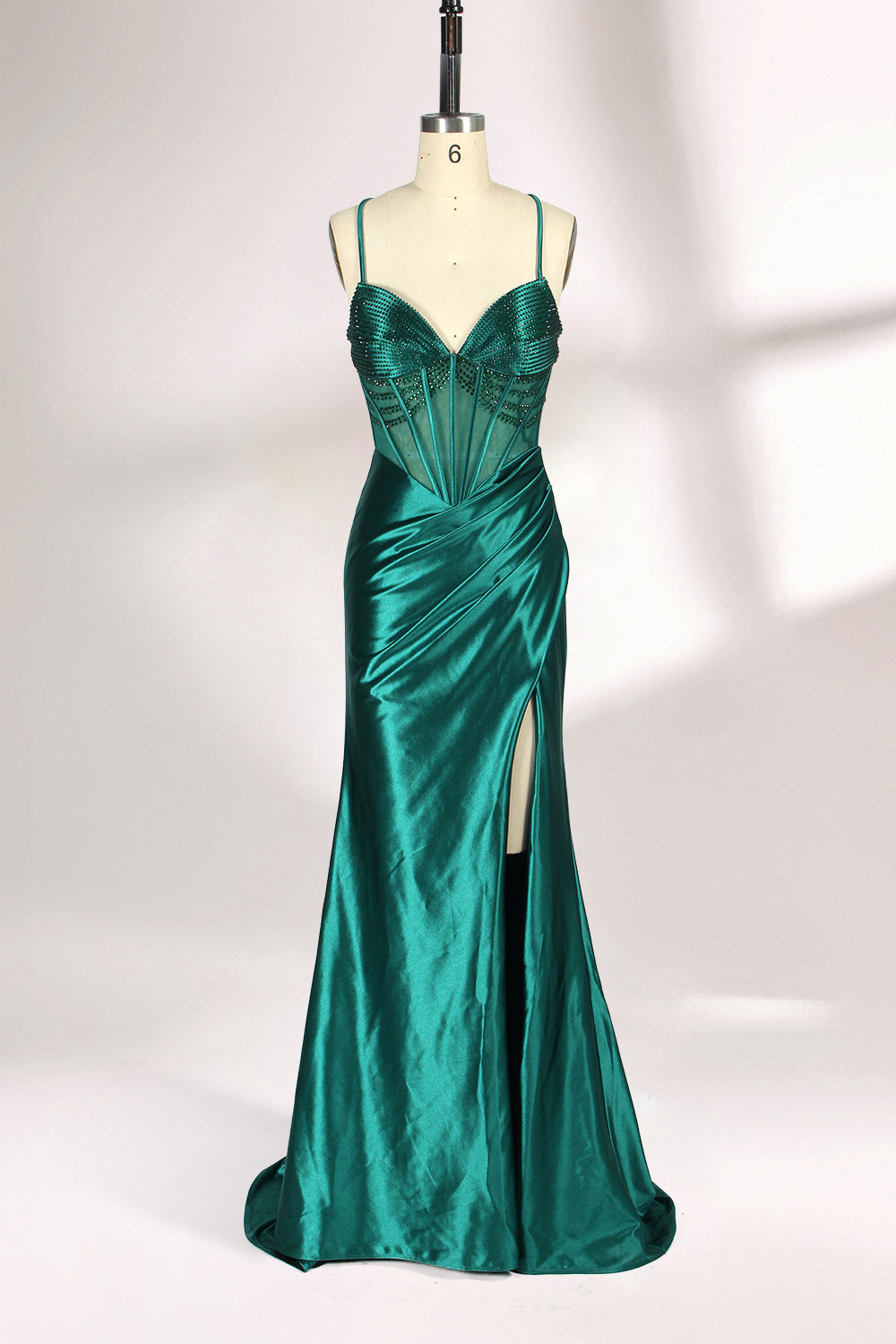 A mannequin displays a green satin gown against a light backdrop. It has thin straps, a sweetheart neckline, and a sheer bodice with beading. The mermaid skirt, featuring a high side slit, combines elegance and allure, perfect for formal galas.1