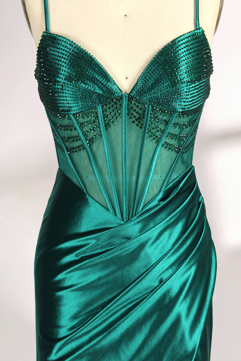 A mannequin displays a green satin gown against a light backdrop. It has thin straps, a sweetheart neckline, and a sheer bodice with beading. The mermaid skirt, featuring a high side slit, combines elegance and allure, perfect for formal galas.4