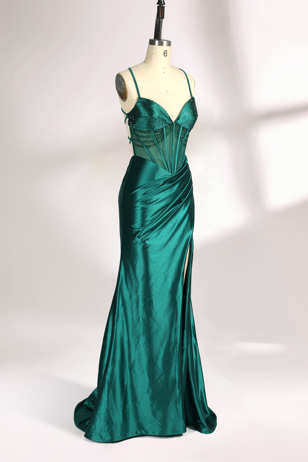 A mannequin displays a green satin gown against a light backdrop. It has thin straps, a sweetheart neckline, and a sheer bodice with beading. The mermaid skirt, featuring a high side slit, combines elegance and allure, perfect for formal galas.3
