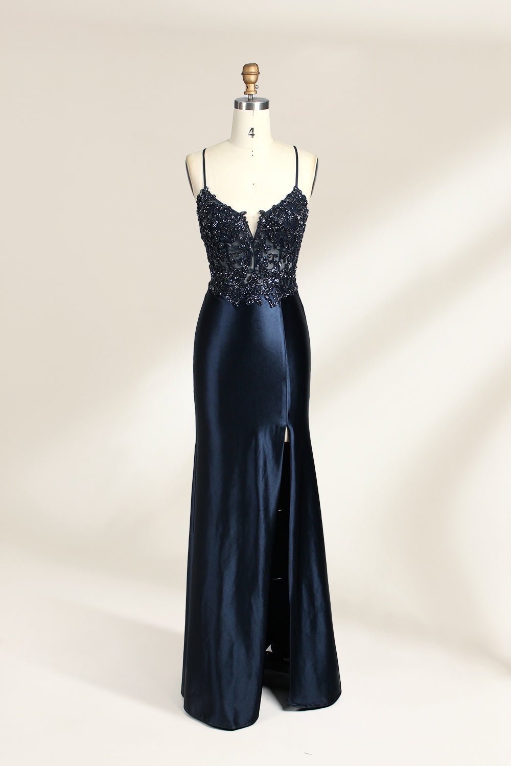 Photograph of a mannequin showcasing a floor-length, navy blue satin evening gown with intricate black sequin embroidery on the bodice and straps. The gown features a deep V-neckline, spaghetti straps, and a high slit on the right side. The background is a plain, light beige wall, enhancing the dress's elegant, sophisticated design.01