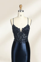 Photograph of a mannequin showcasing a floor-length, navy blue satin evening gown with intricate black sequin embroidery on the bodice and straps. The gown features a deep V-neckline, spaghetti straps, and a high slit on the right side. The background is a plain, light beige wall, enhancing the dress's elegant, sophisticated design.03