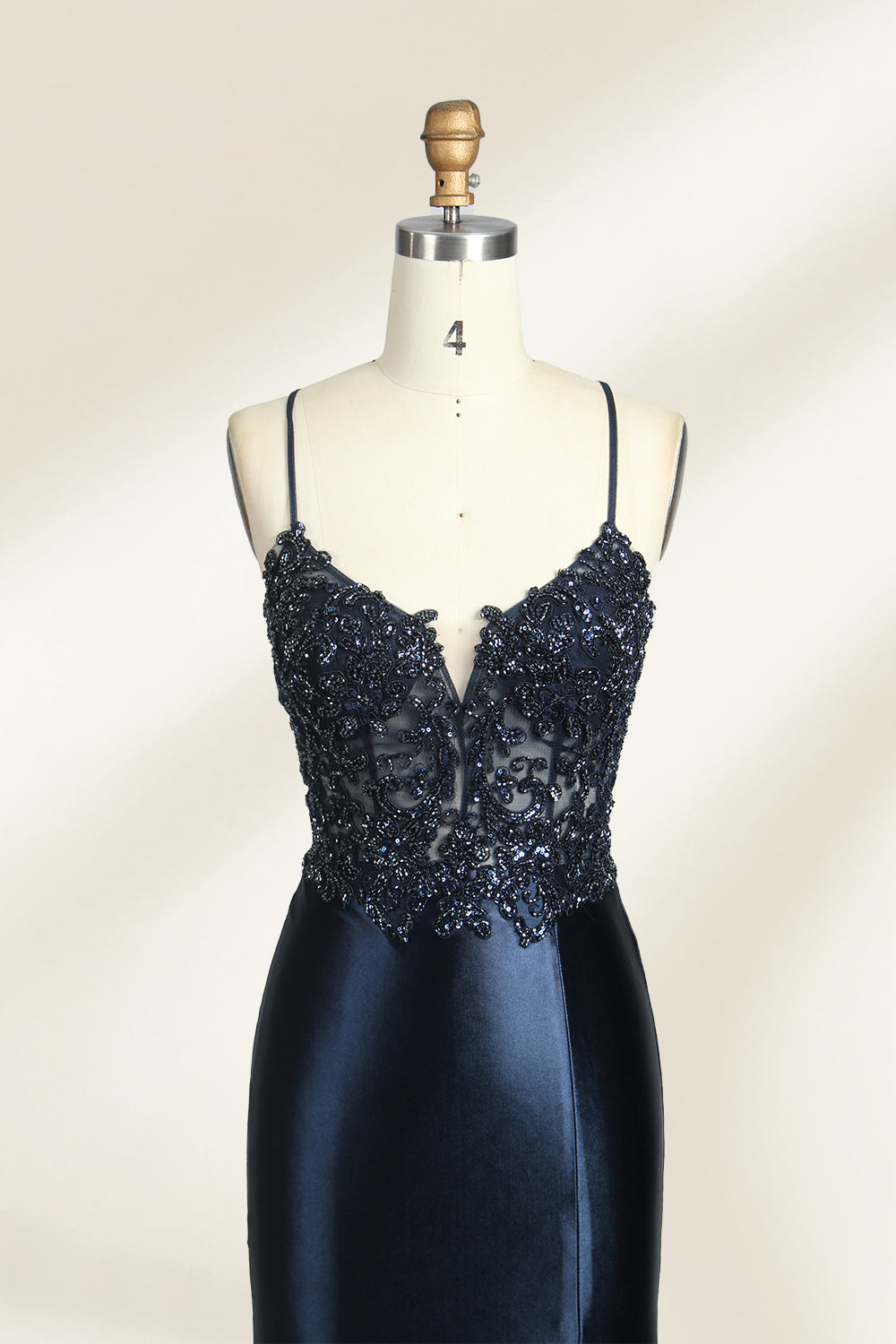 Photograph of a mannequin showcasing a floor-length, navy blue satin evening gown with intricate black sequin embroidery on the bodice and straps. The gown features a deep V-neckline, spaghetti straps, and a high slit on the right side. The background is a plain, light beige wall, enhancing the dress's elegant, sophisticated design.03
