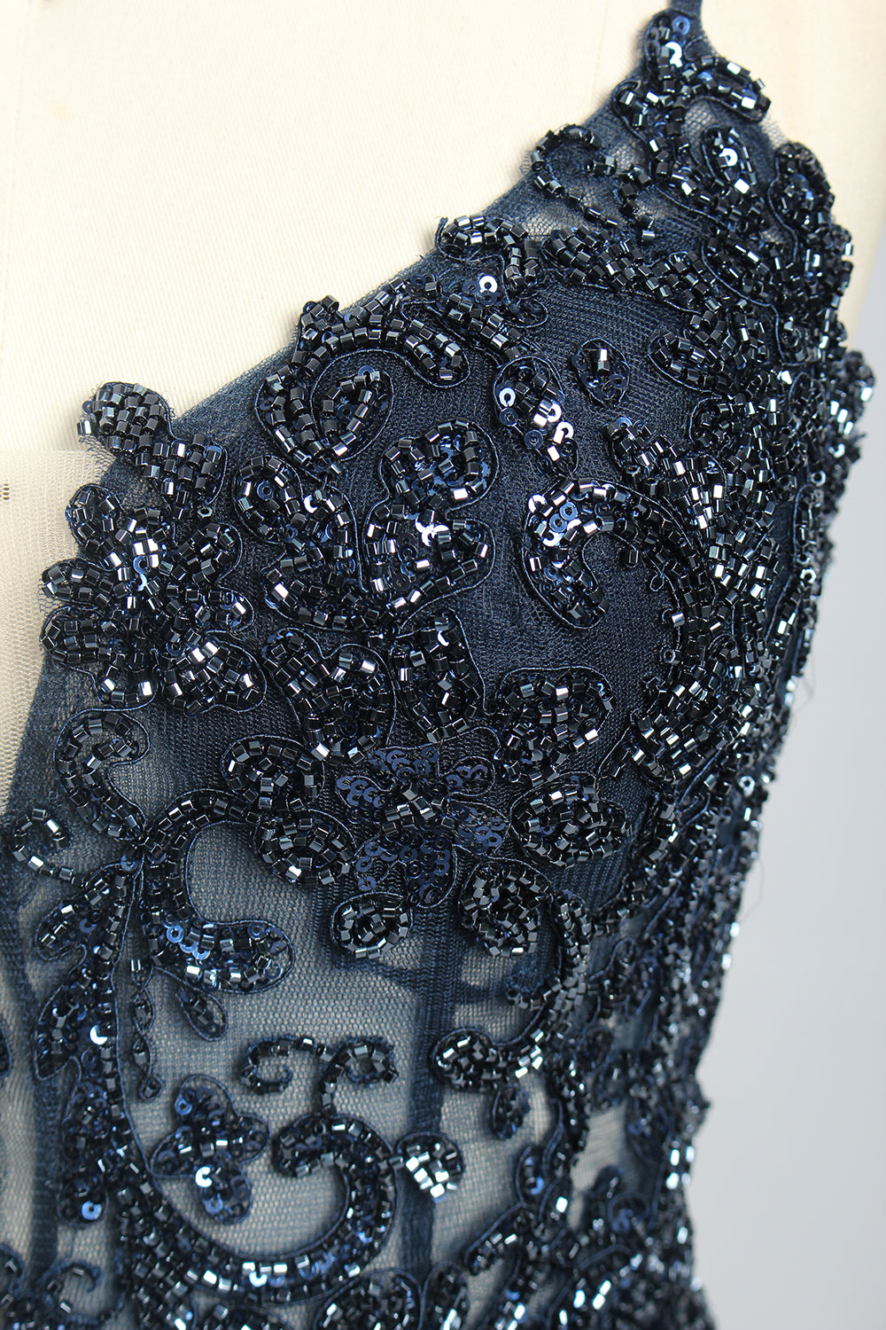 Photograph of a mannequin showcasing a floor-length, navy blue satin evening gown with intricate black sequin embroidery on the bodice and straps. The gown features a deep V-neckline, spaghetti straps, and a high slit on the right side. The background is a plain, light beige wall, enhancing the dress's elegant, sophisticated design.06