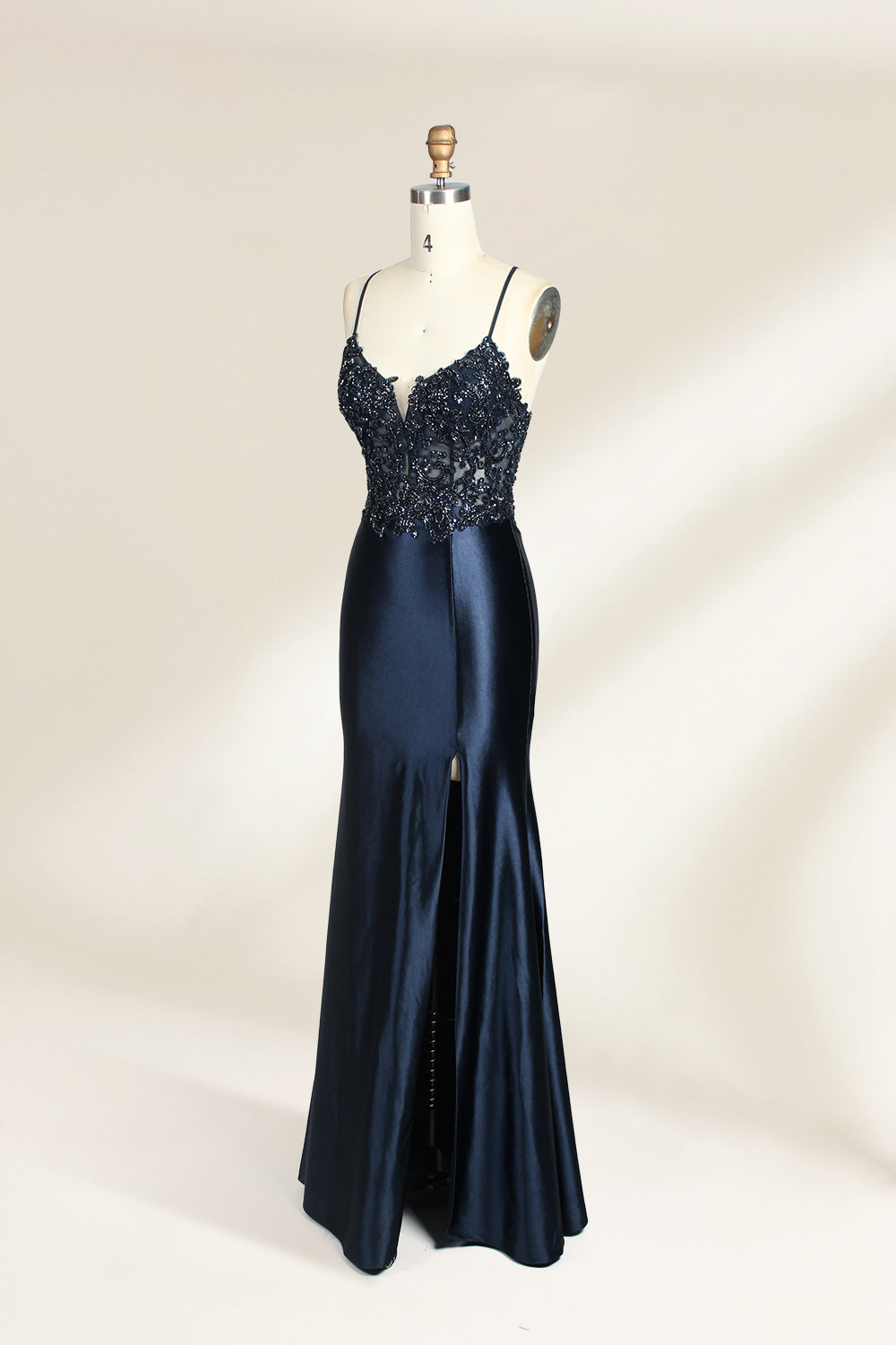 Photograph of a mannequin showcasing a floor-length, navy blue satin evening gown with intricate black sequin embroidery on the bodice and straps. The gown features a deep V-neckline, spaghetti straps, and a high slit on the right side. The background is a plain, light beige wall, enhancing the dress's elegant, sophisticated design.04