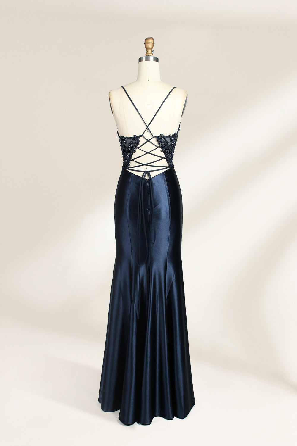 Photograph of a mannequin showcasing a floor-length, navy blue satin evening gown with intricate black sequin embroidery on the bodice and straps. The gown features a deep V-neckline, spaghetti straps, and a high slit on the right side. The background is a plain, light beige wall, enhancing the dress's elegant, sophisticated design.02