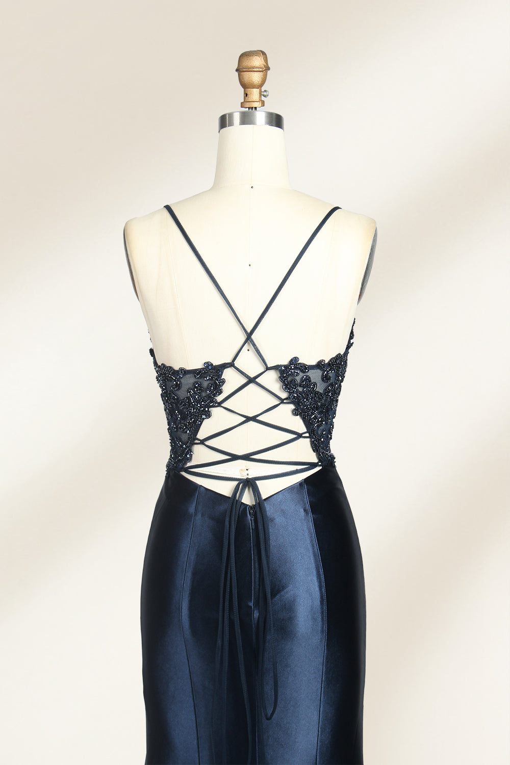 Photograph of a mannequin showcasing a floor-length, navy blue satin evening gown with intricate black sequin embroidery on the bodice and straps. The gown features a deep V-neckline, spaghetti straps, and a high slit on the right side. The background is a plain, light beige wall, enhancing the dress's elegant, sophisticated design.05