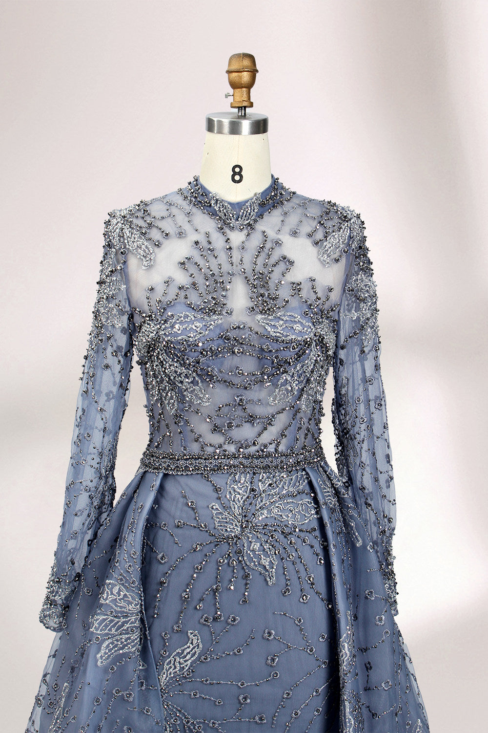 Presented on a mannequin is a blue - toned evening gown. It has a high neckline, long sheer sleeves, and intricate floral embroidery. The elegant design gives off a luxurious and sophisticated vibe, ideal for formal occasions.7