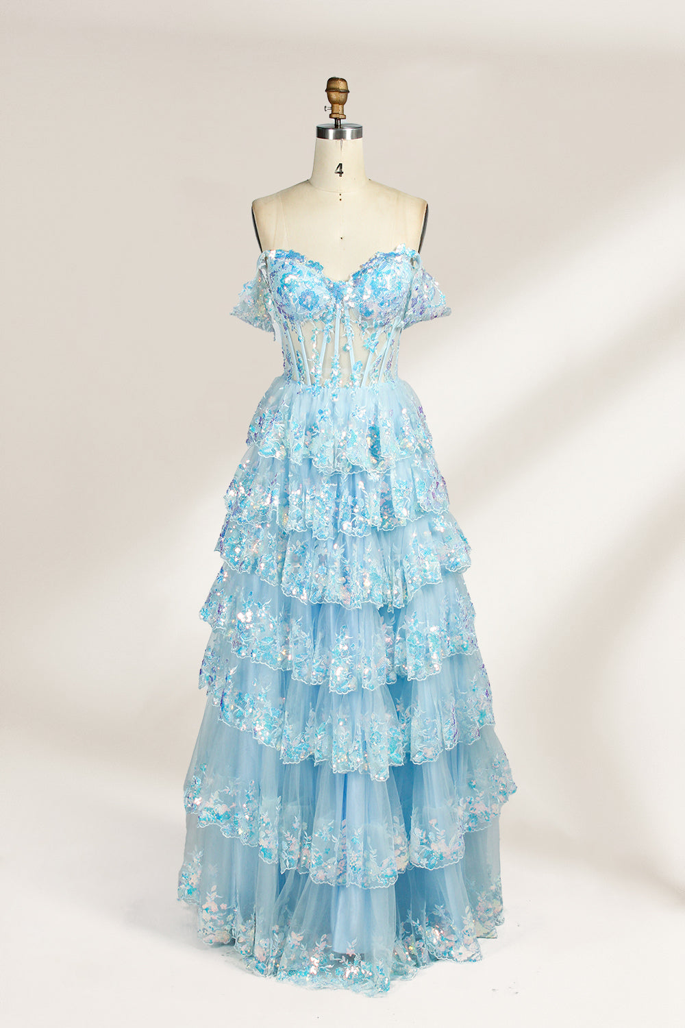 Photograph of a mannequin displaying a strapless, multi-layered, light blue tulle gown adorned with intricate floral embroidery. The gown has an off-shoulder neckline and a fitted bodice, cascading into voluminous, ruffled tiers. The background is plain white, highlighting the dress's delicate details. The mannequin is marked with the number "4" on its torso.1