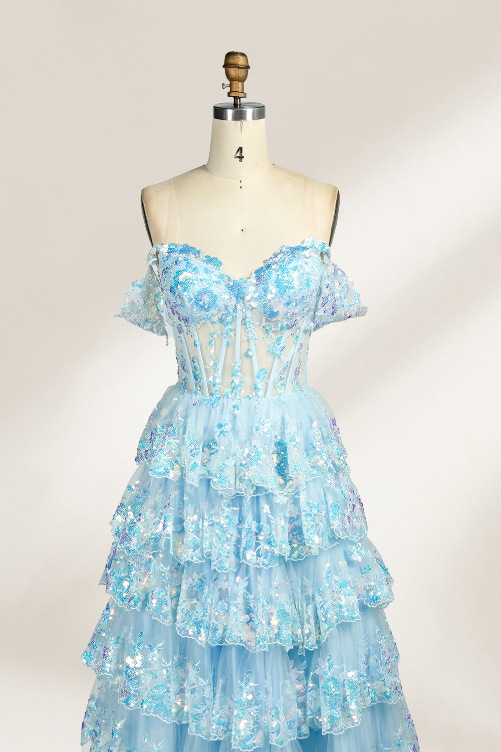 Photograph of a mannequin displaying a strapless, multi-layered, light blue tulle gown adorned with intricate floral embroidery. The gown has an off-shoulder neckline and a fitted bodice, cascading into voluminous, ruffled tiers. The background is plain white, highlighting the dress's delicate details. The mannequin is marked with the number "4" on its torso.5
