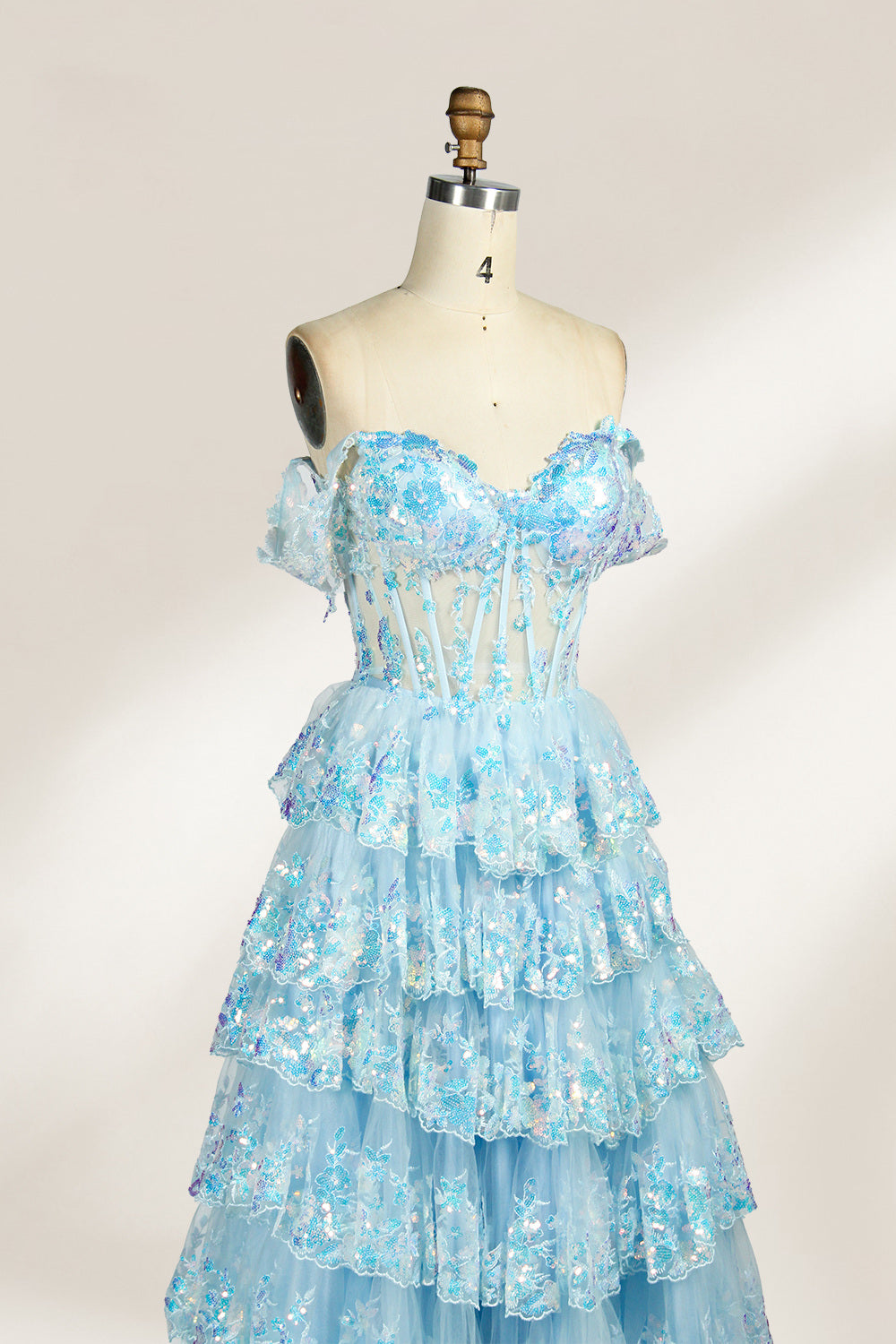 Photograph of a mannequin displaying a strapless, multi-layered, light blue tulle gown adorned with intricate floral embroidery. The gown has an off-shoulder neckline and a fitted bodice, cascading into voluminous, ruffled tiers. The background is plain white, highlighting the dress's delicate details. The mannequin is marked with the number "4" on its torso.4