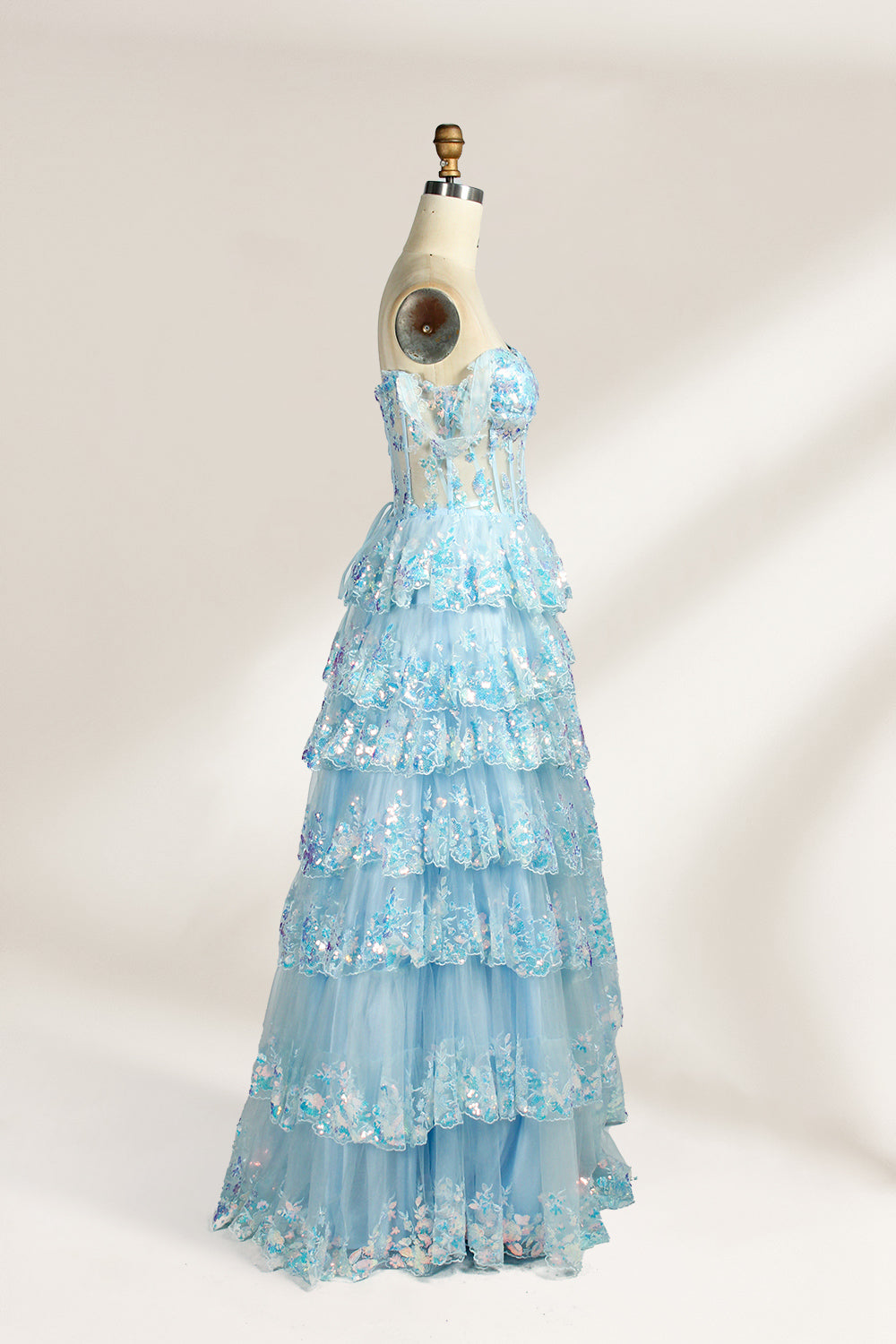 Photograph of a mannequin displaying a strapless, multi-layered, light blue tulle gown adorned with intricate floral embroidery. The gown has an off-shoulder neckline and a fitted bodice, cascading into voluminous, ruffled tiers. The background is plain white, highlighting the dress's delicate details. The mannequin is marked with the number "4" on its torso.