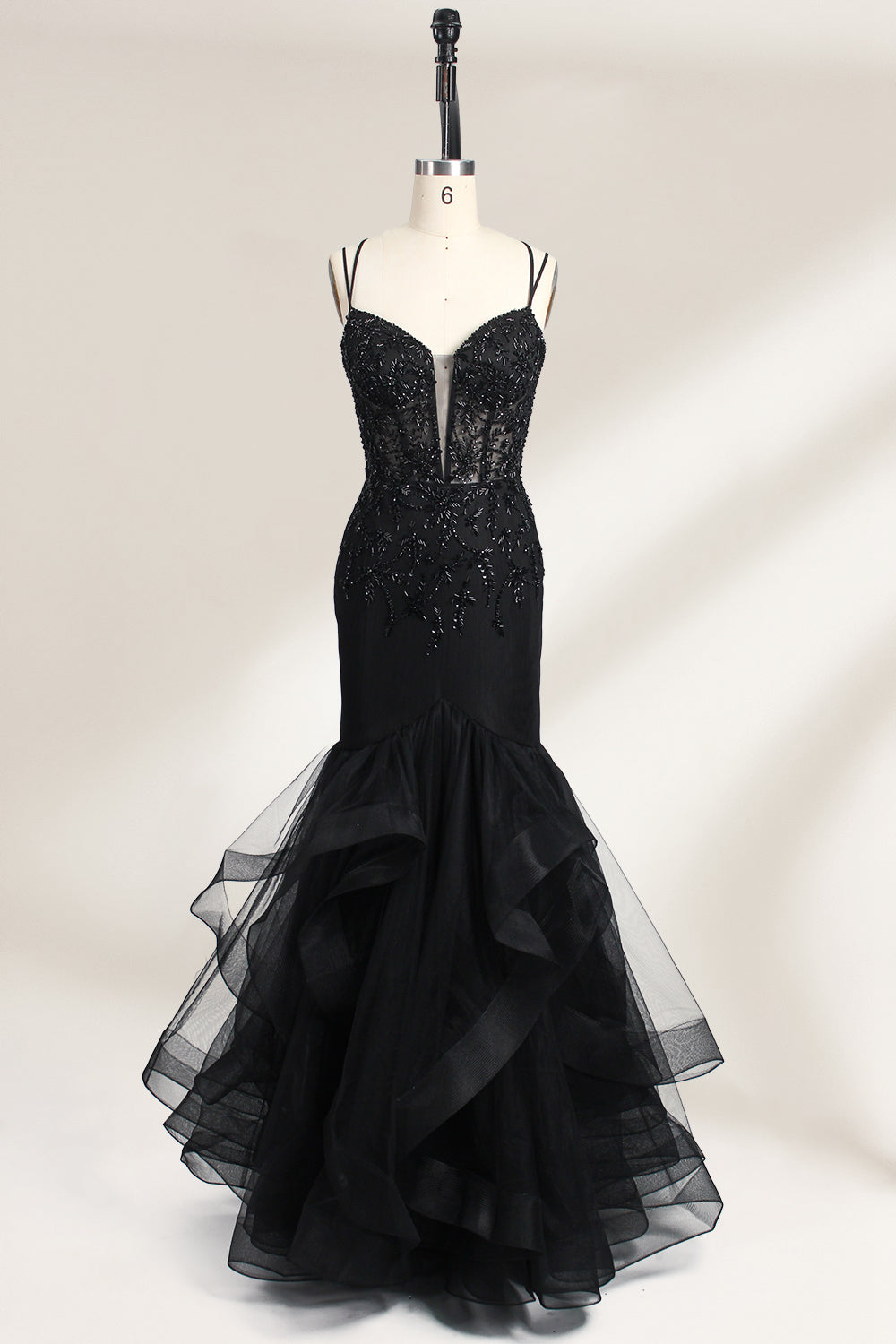 A mannequin displays a black mermaid - style gown. It has thin straps, a deep V - neckline with sheer panels, and intricate black bead embroidery. The layered tulle skirt adds a romantic touch, perfect for formal events like galas or weddings, exuding elegance and sophistication.1