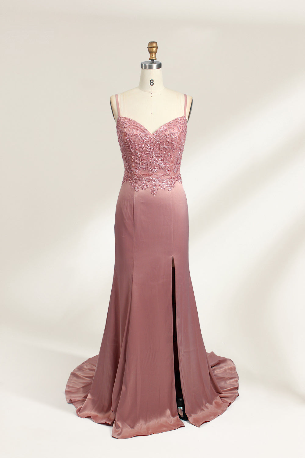 A photograph of a model mannequin wearing a floor-length, mauve satin gown with a deep V-neckline. The bodice is adorned with intricate lace detailing, featuring floral patterns and a subtle sheen. The gown has a thigh-high slit on the right side, adding a touch of elegance. The mannequin stands against a plain white background, highlighting the dress's smooth texture and elegant silhouette.01