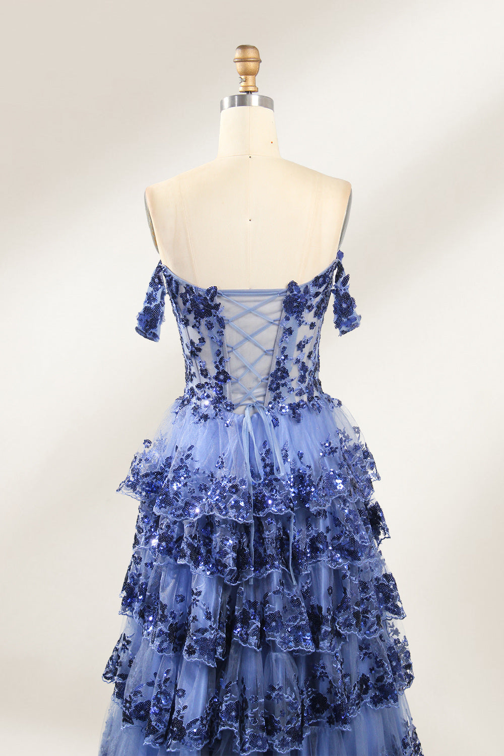 Photograph of a mannequin displaying a strapless, multi-layered, light blue tulle gown adorned with intricate floral embroidery. The gown has an off-shoulder neckline and a fitted bodice, cascading into voluminous, ruffled tiers. The background is plain white, highlighting the dress's delicate details. The mannequin is marked with the number "4" on its torso.6