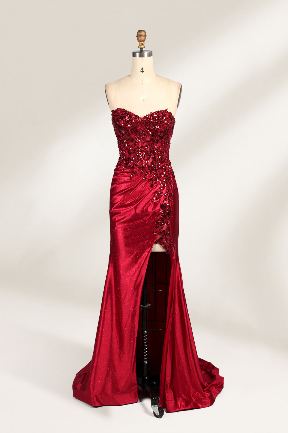 This photograph features a mannequin dressed in a strapless, floor-length, deep red satin evening gown. The gown is adorned with intricate sequin embroidery on the bodice and a high slit on the left side, revealing black stockings. The fabric has a smooth, luxurious texture. The mannequin stands on a black stool against a plain white background, creating a stark contrast that highlights the gown's vibrant color and detailed design. The overall style is elegant and glamorous, suitable for formal occasions.1