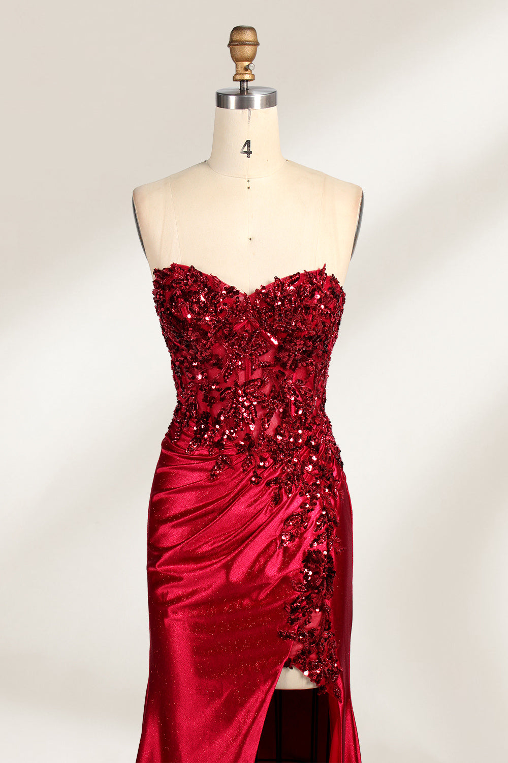 This photograph features a mannequin dressed in a strapless, floor-length, deep red satin evening gown. The gown is adorned with intricate sequin embroidery on the bodice and a high slit on the left side, revealing black stockings. The fabric has a smooth, luxurious texture. The mannequin stands on a black stool against a plain white background, creating a stark contrast that highlights the gown's vibrant color and detailed design. The overall style is elegant and glamorous, suitable for formal occasions.3