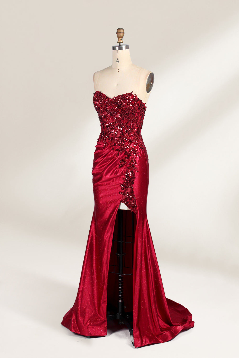 This photograph features a mannequin dressed in a strapless, floor-length, deep red satin evening gown. The gown is adorned with intricate sequin embroidery on the bodice and a high slit on the left side, revealing black stockings. The fabric has a smooth, luxurious texture. The mannequin stands on a black stool against a plain white background, creating a stark contrast that highlights the gown's vibrant color and detailed design. The overall style is elegant and glamorous, suitable for formal occasions.4