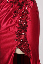 This photograph features a mannequin dressed in a strapless, floor-length, deep red satin evening gown. The gown is adorned with intricate sequin embroidery on the bodice and a high slit on the left side, revealing black stockings. The fabric has a smooth, luxurious texture. The mannequin stands on a black stool against a plain white background, creating a stark contrast that highlights the gown's vibrant color and detailed design. The overall style is elegant and glamorous, suitable for formal occasions.4