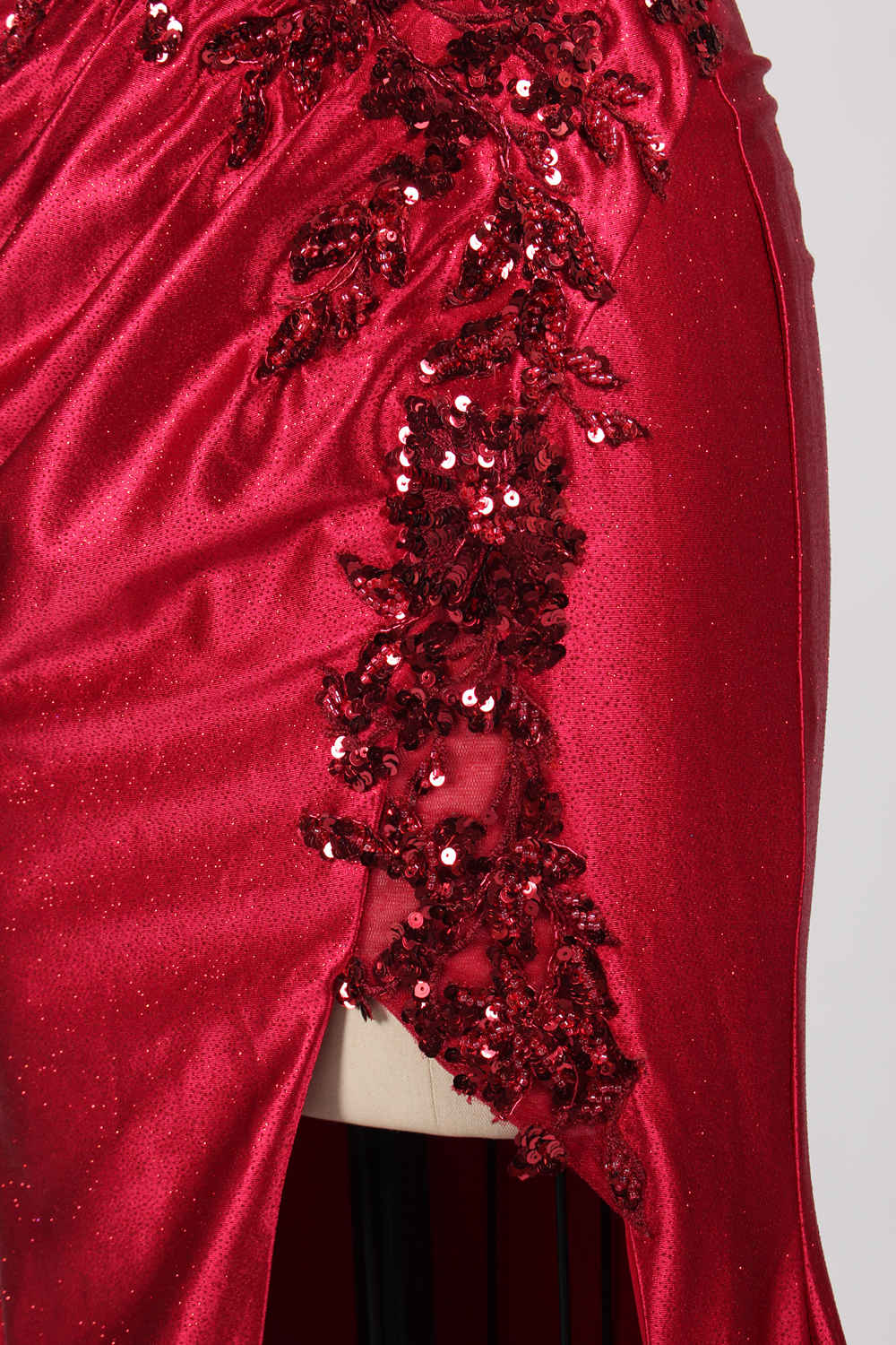 This photograph features a mannequin dressed in a strapless, floor-length, deep red satin evening gown. The gown is adorned with intricate sequin embroidery on the bodice and a high slit on the left side, revealing black stockings. The fabric has a smooth, luxurious texture. The mannequin stands on a black stool against a plain white background, creating a stark contrast that highlights the gown's vibrant color and detailed design. The overall style is elegant and glamorous, suitable for formal occasions.4