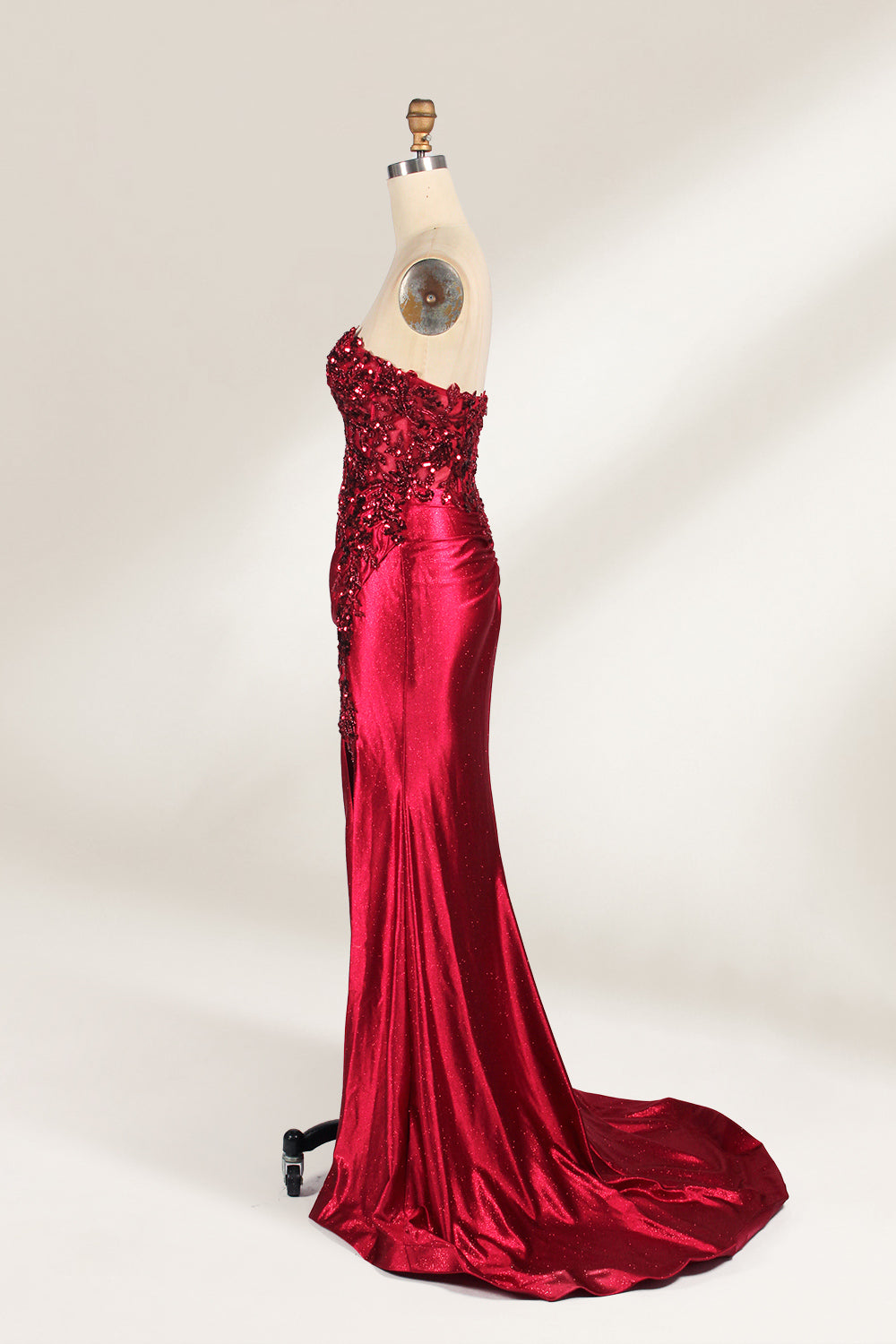 This photograph features a mannequin dressed in a strapless, floor-length, deep red satin evening gown. The gown is adorned with intricate sequin embroidery on the bodice and a high slit on the left side, revealing black stockings. The fabric has a smooth, luxurious texture. The mannequin stands on a black stool against a plain white background, creating a stark contrast that highlights the gown's vibrant color and detailed design. The overall style is elegant and glamorous, suitable for formal occasions.5