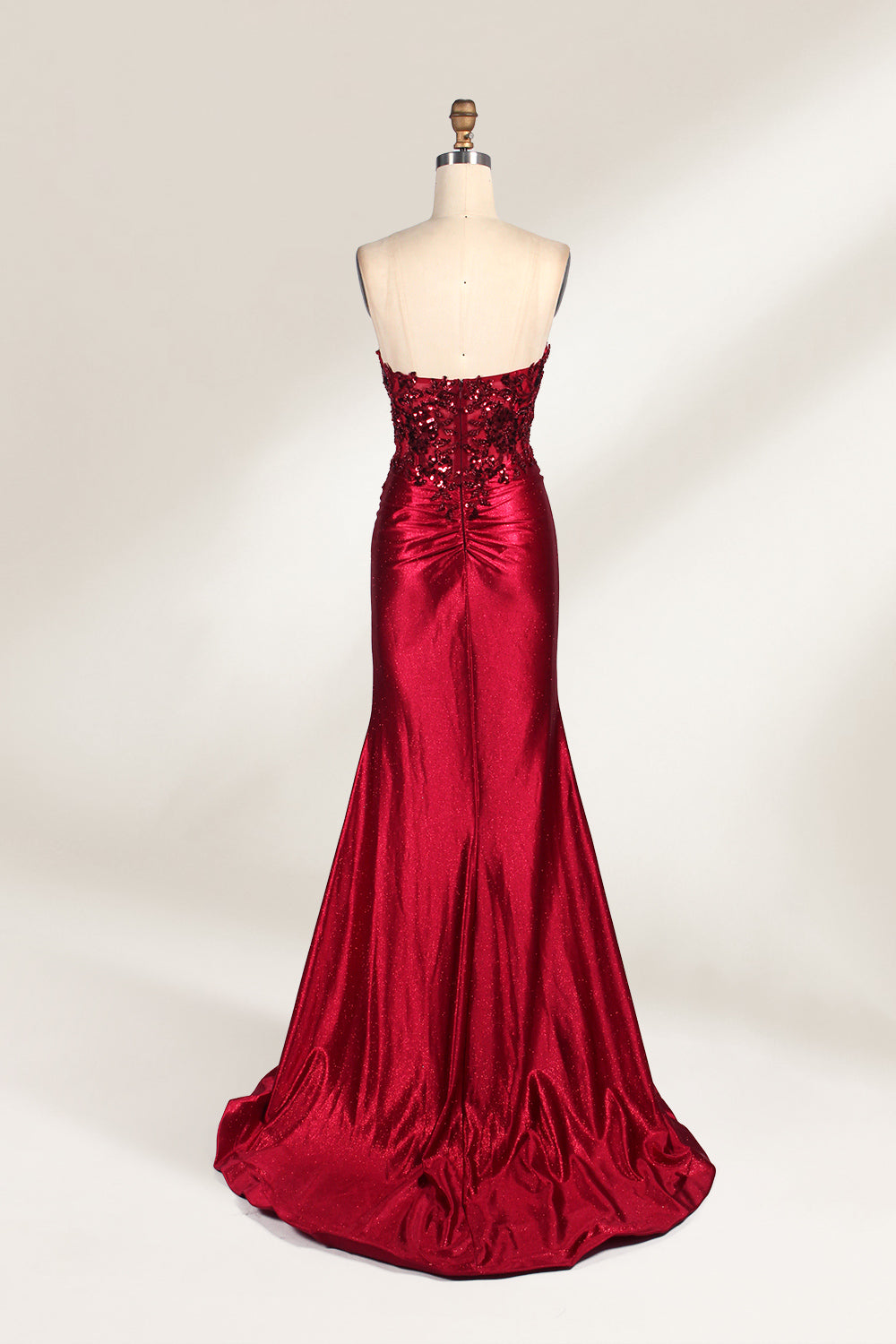 This photograph features a mannequin dressed in a strapless, floor-length, deep red satin evening gown. The gown is adorned with intricate sequin embroidery on the bodice and a high slit on the left side, revealing black stockings. The fabric has a smooth, luxurious texture. The mannequin stands on a black stool against a plain white background, creating a stark contrast that highlights the gown's vibrant color and detailed design. The overall style is elegant and glamorous, suitable for formal occasions.2